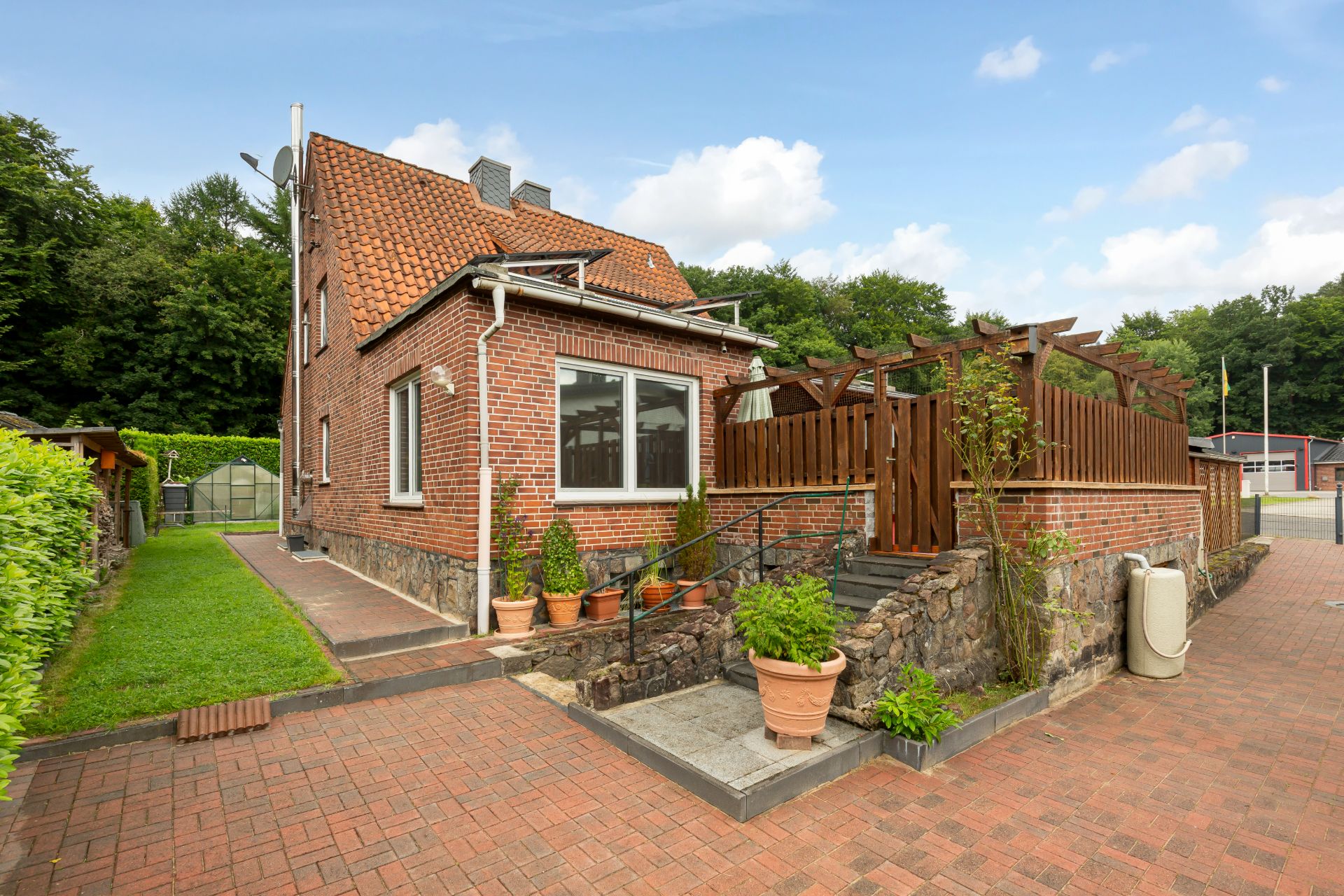 Backsteinwohnhaus mit rotem Dach, Terrasse aus Holz und mehreren Pflanzkübeln im Eingangsbereich.