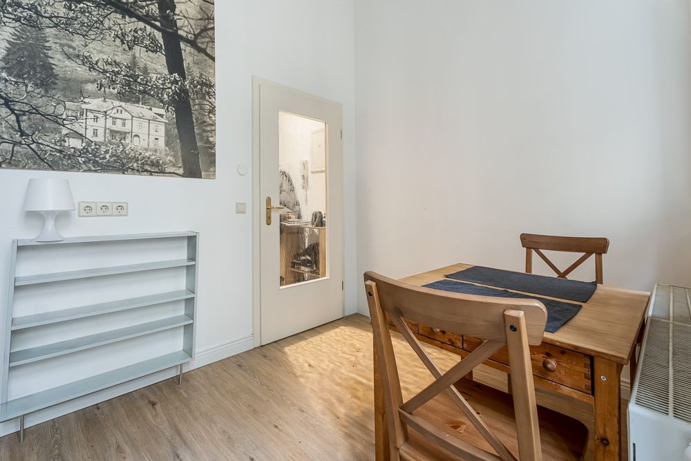 Kleiner Essbereich mit Holztisch, zwei Stühlen, Regal, Lampe und Wandbild in hellem Raum.