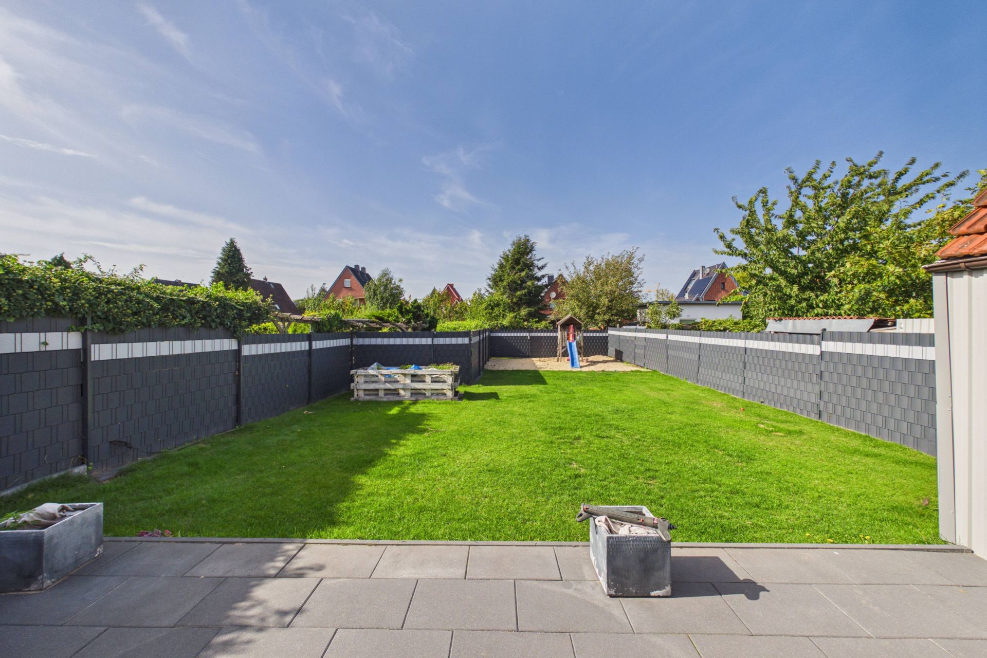 Großer, gepflegter Garten mit Rasen, Spielgerät im Hintergrund und grauem Zaun.