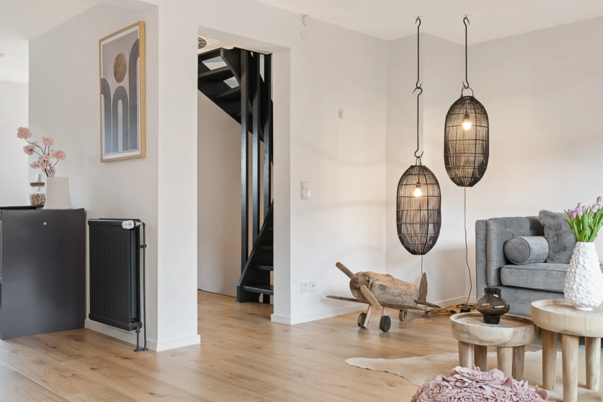 Modernes Wohnzimmer mit grauem Sofa, Holztischen und hängenden Lampen, Treppe im Hintergrund.