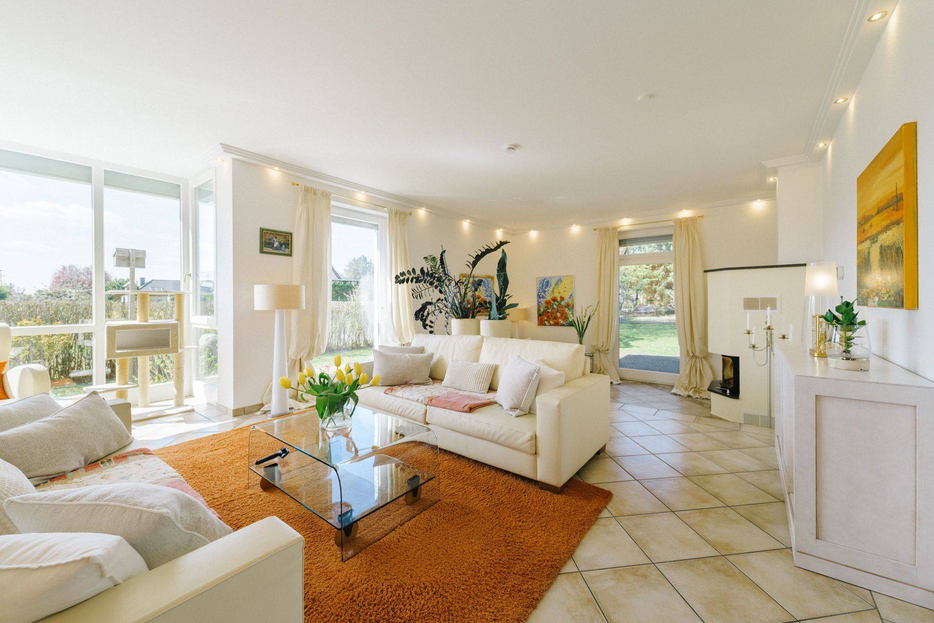 Helles Wohnzimmer mit weißen Sofas, Glas-Couchtisch, orangenem Teppich und großen Fenstern zum Garten.