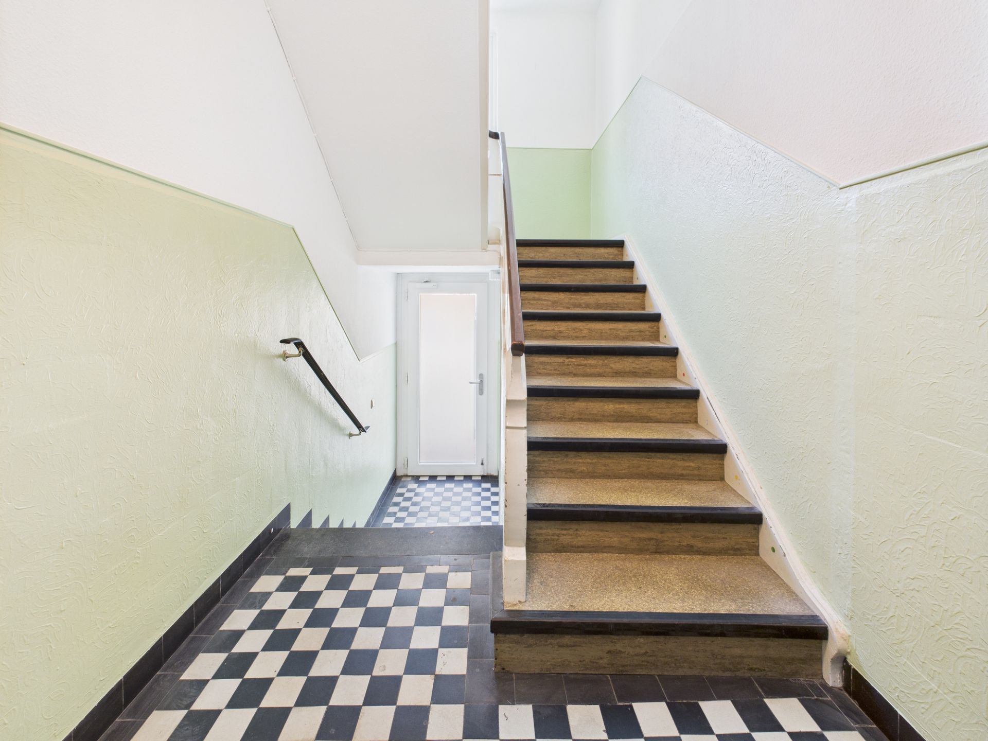 Treppenhaus mit schwarz-weiß kariertem Fliesenboden und grün gestrichenen Wänden.