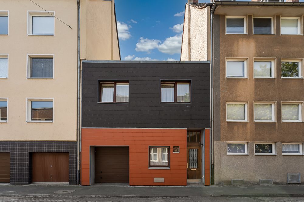 Modernes zweistöckiges Wohnhaus mit dunkler Fassade und orangefarbenen Akzenten zwischen zwei Mehrfamilienhäusern.