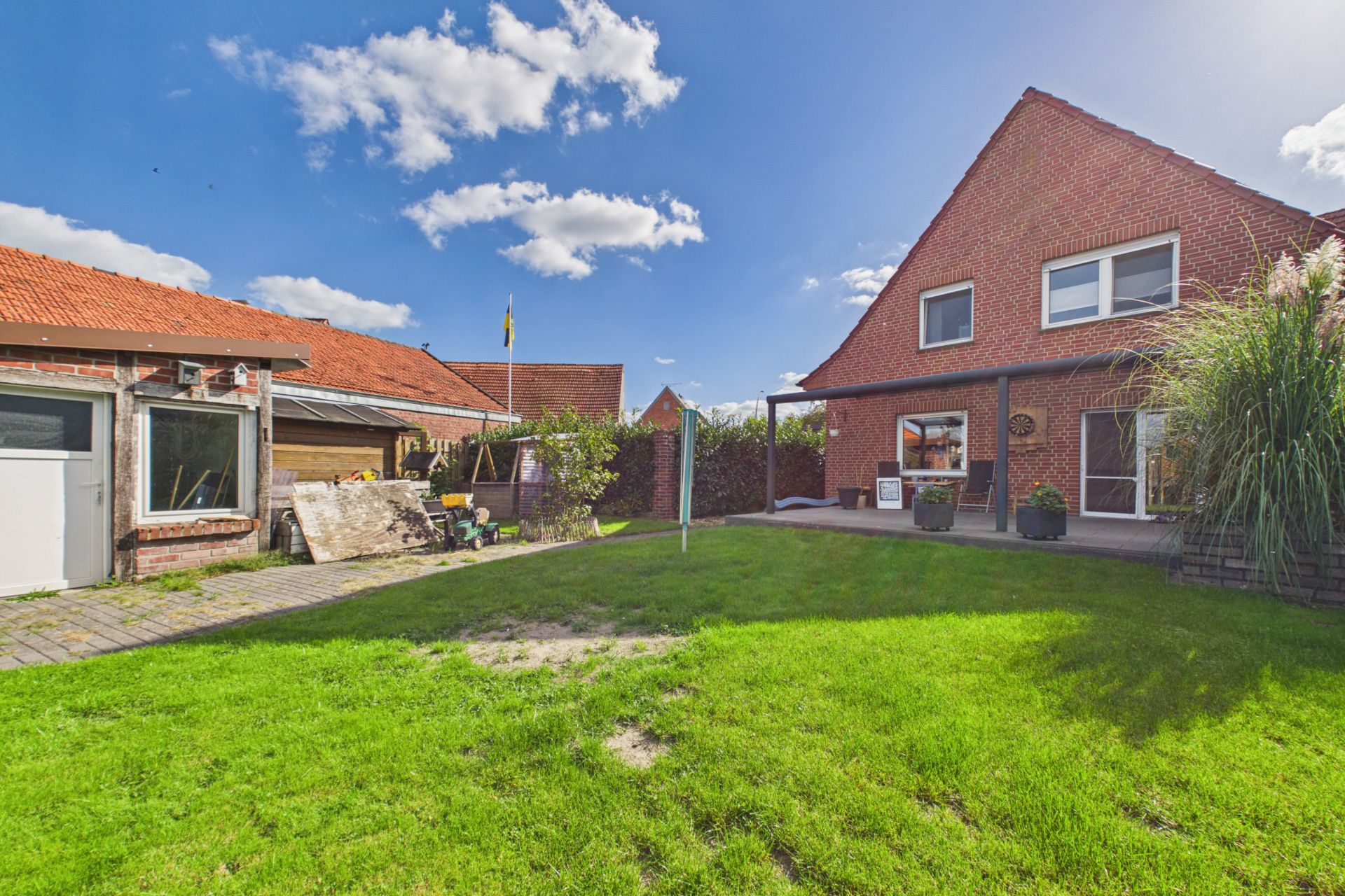 Grüner Garten mit Rasen, Terrasse, Backsteinhaus und Nebengebäude unter blauem Himmel.