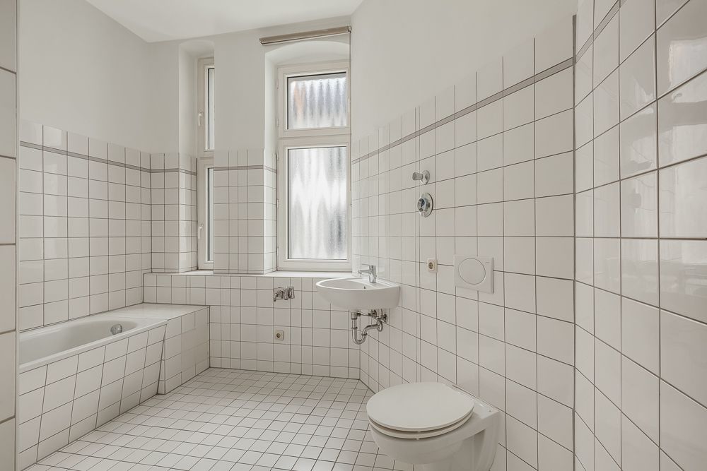 Helles Badezimmer mit Badewanne, Waschbecken, Toilette und zwei Fenstern, alles weiß gefliest.