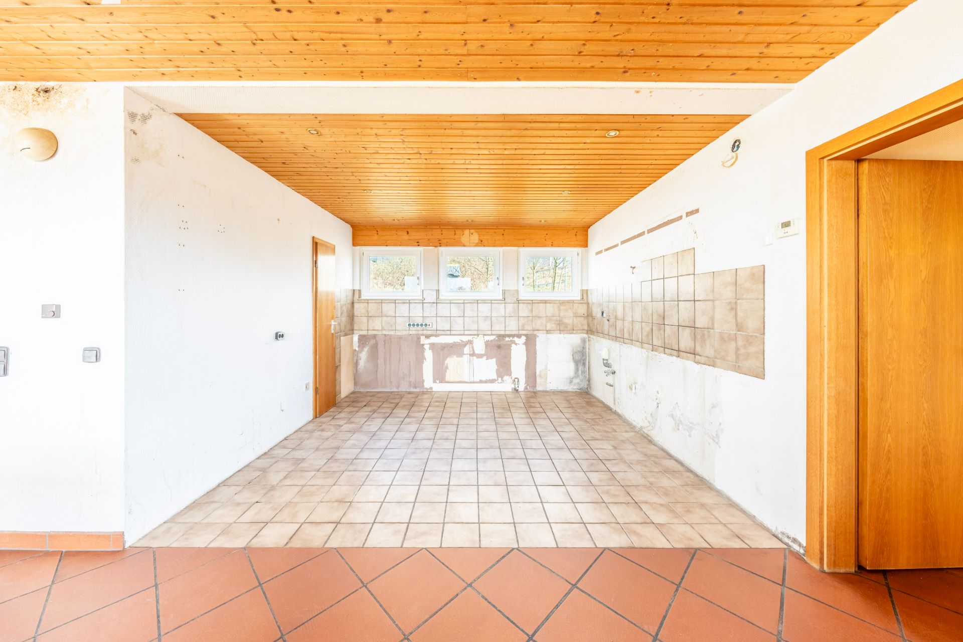 Leerer Raum mit gefliestem Boden, Holzdecke und drei Fenstern an der Rückwand.