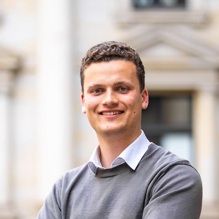 Ein junger Mann steht draußen mit verschränkten Armen und lächelt in die Kamera.