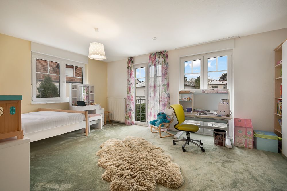 Helles Kinderzimmer mit Bett, Schreibtisch, Regal und großem Fenster.