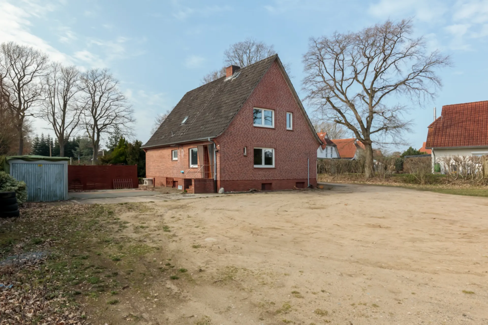 Backstein-Einfamilienhaus mit Satteldach auf großem, unbefestigtem Hof und Bäumen im Hintergrund.