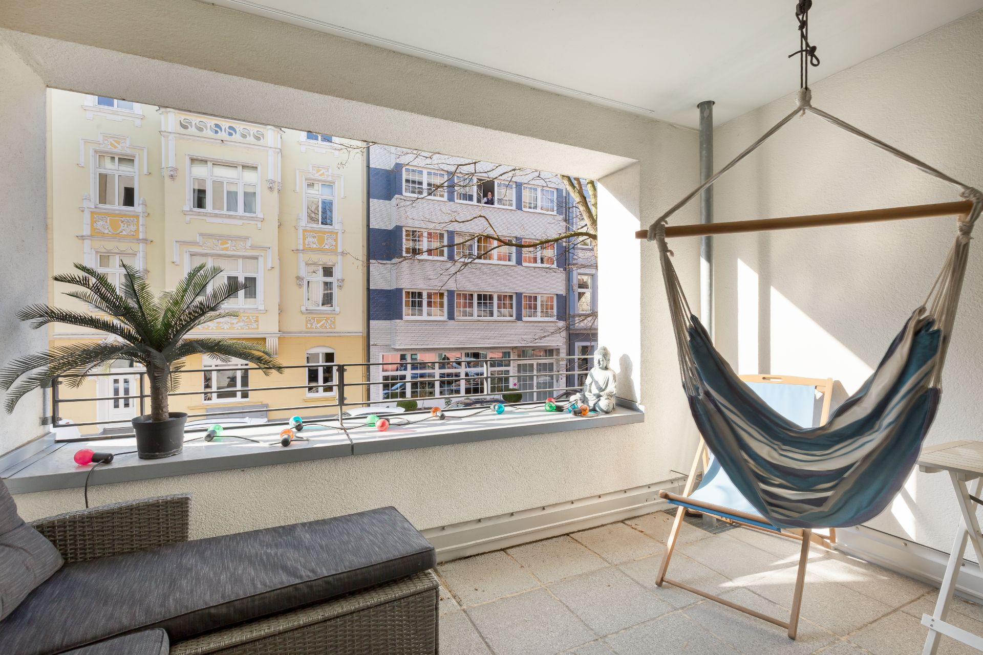 Überdachter Balkon mit Hängesessel, Sessel, Pflanze und Blick auf bunte Wohnhäuser.