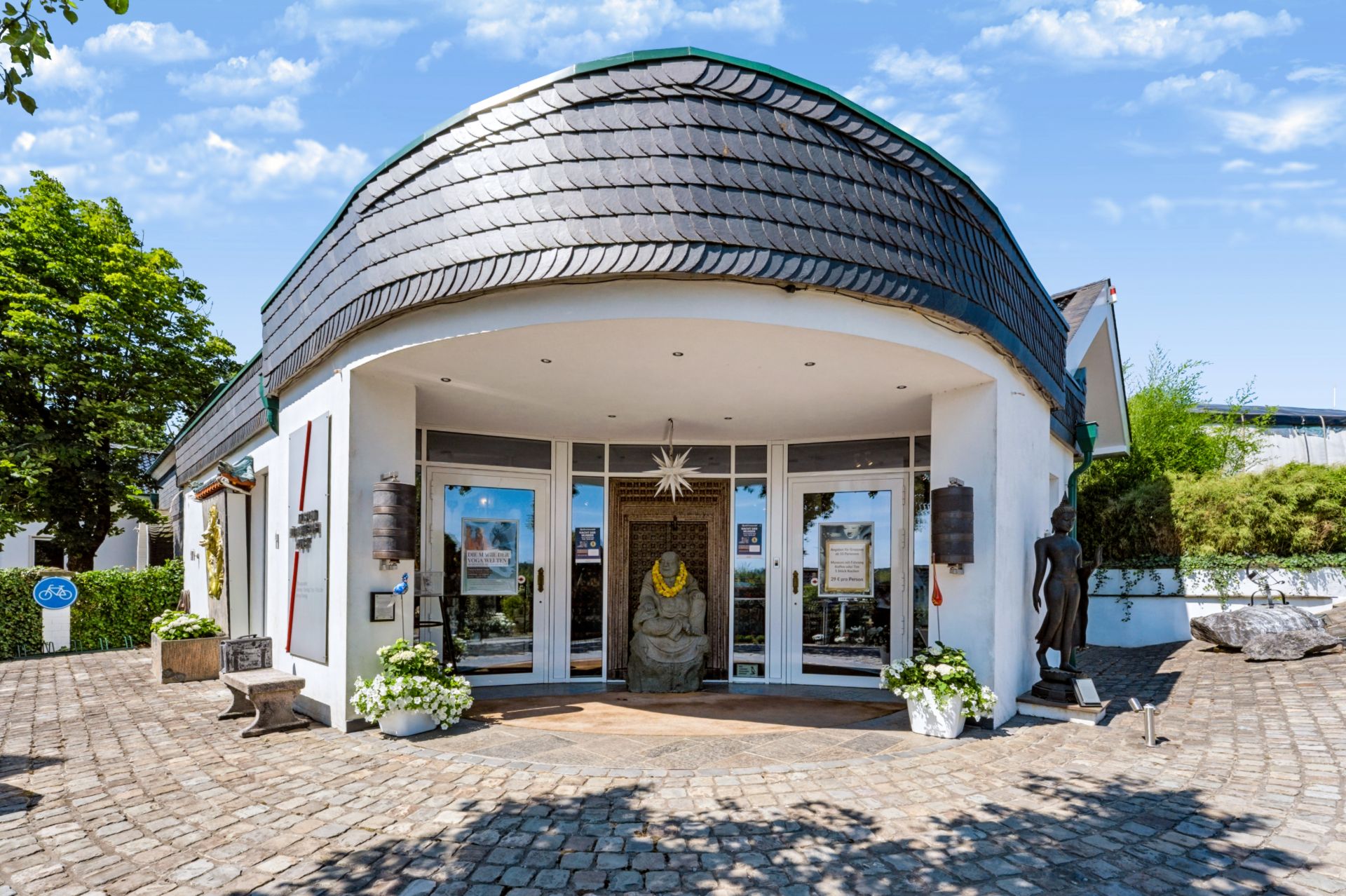 Runder Eingangsbereich mit Glasfront, Pflanzkübeln, Statue und Kopfsteinpflaster.