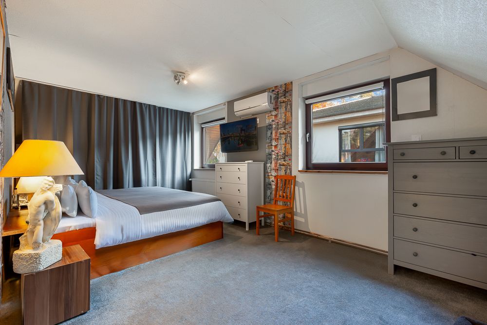 Helles Schlafzimmer mit Doppelbett, grauen Vorhängen, Kommoden, Stuhl und Lampe auf einem Nachttisch.