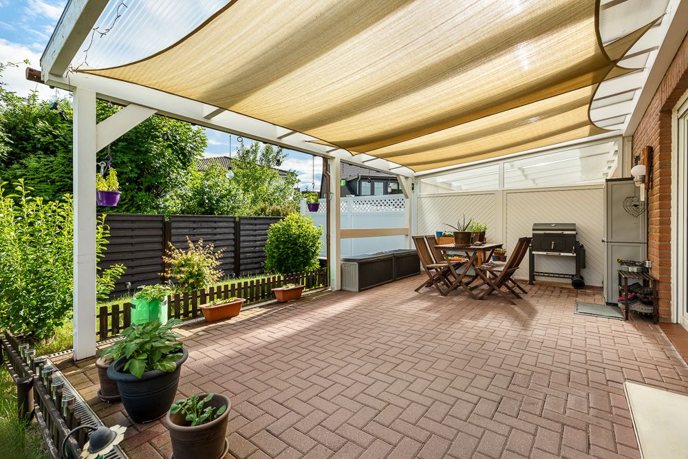 Überdachte Terrasse mit Sitzgruppe, Grill und Blumentöpfen, daneben ein Holzzaun und viele Pflanzen.
