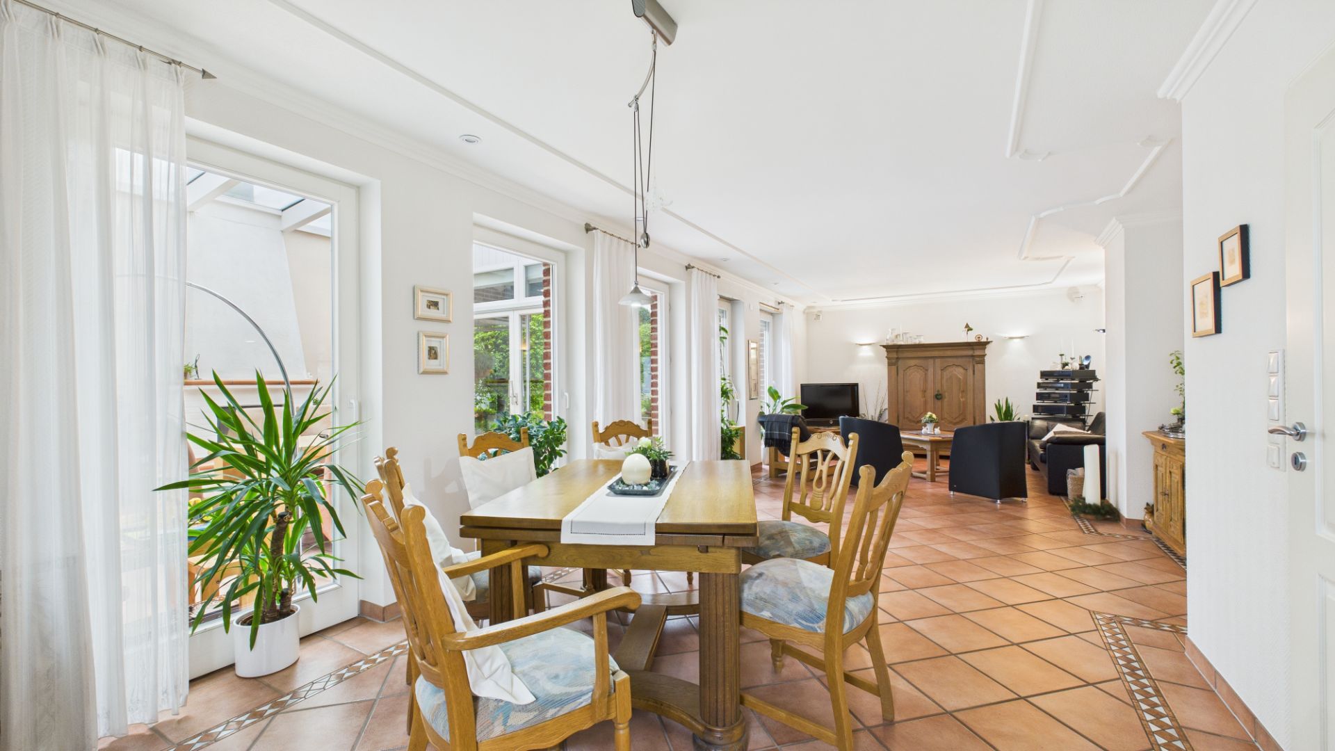 Helles Wohnzimmer mit Holztisch, sechs Stühlen, Pflanzen und großen Fenstern.