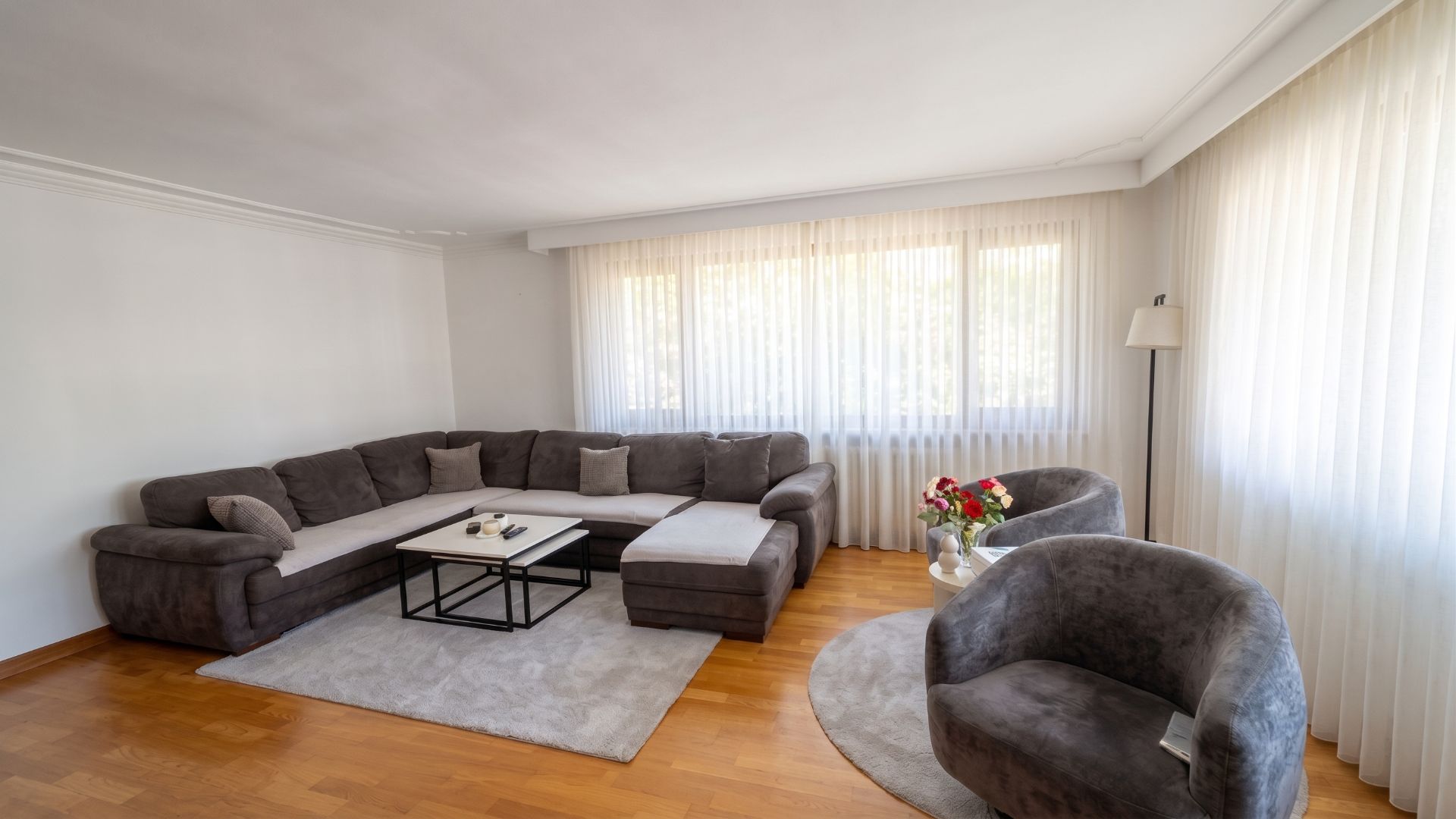 Wohnzimmer mit grauem Ecksofa, zwei Sesseln, Couchtisch und großen Fenstern mit Vorhängen.