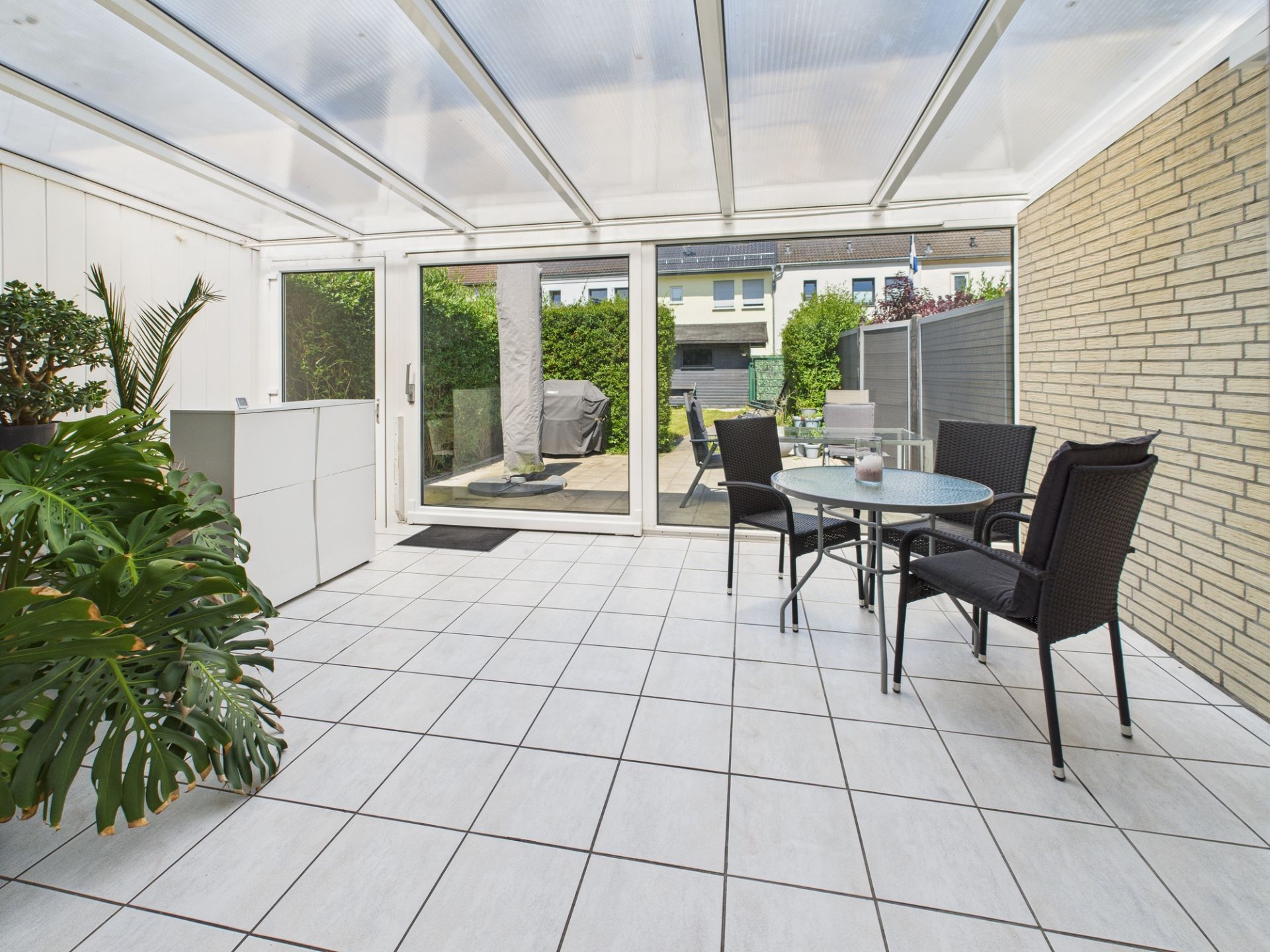 Überdachte Terrasse mit Glastür, Tisch und vier Stühlen, Pflanzen in der Ecke.