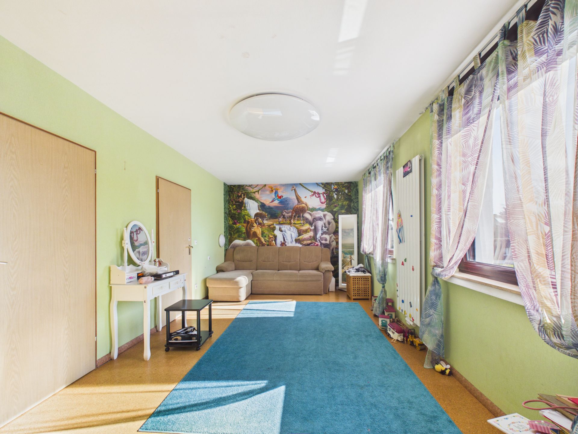 Helles Wohnzimmer mit Sofa, blauem Teppich und Tier-Wandbild, große Fenster mit bunten Vorhängen.
