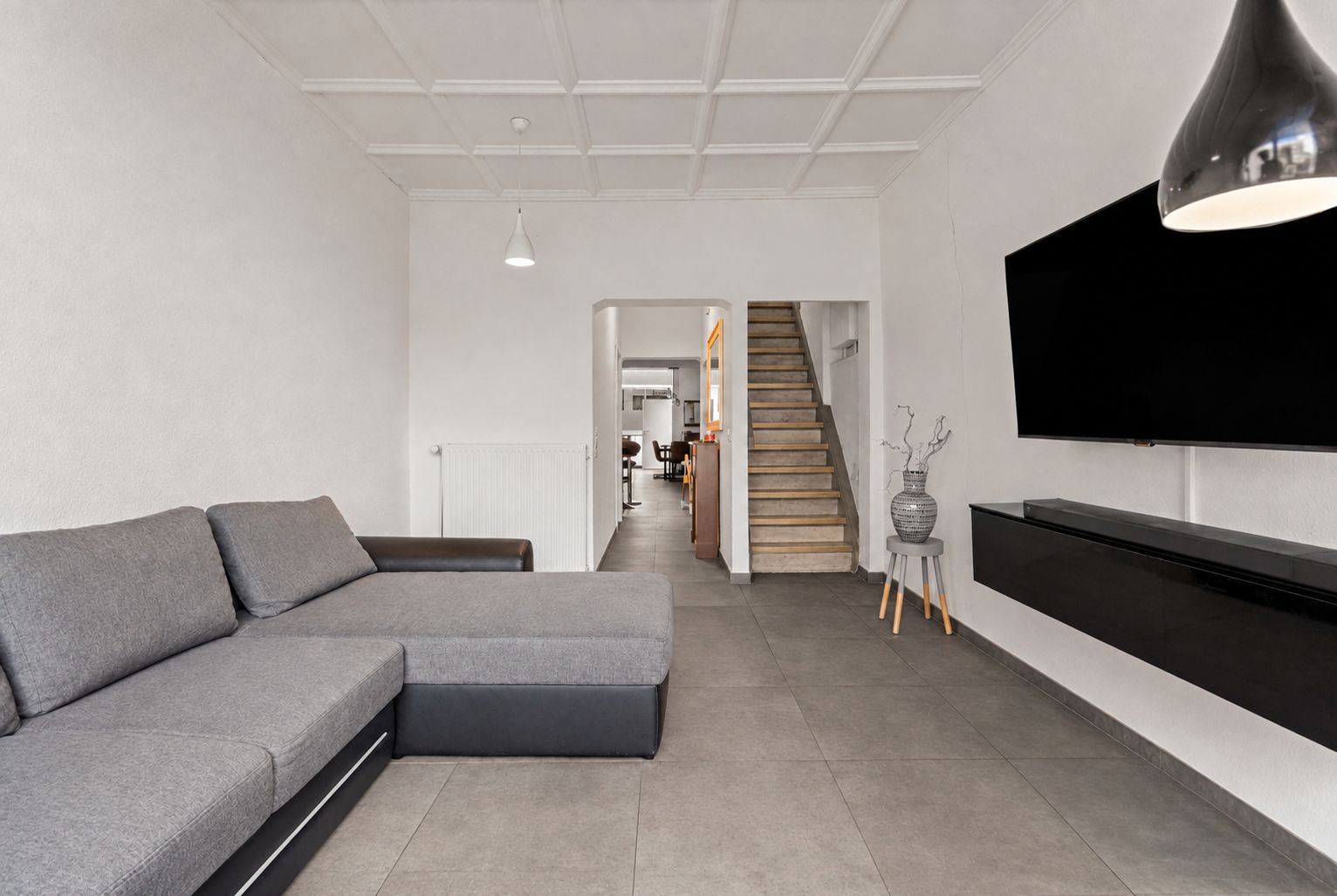 Modernes Wohnzimmer mit grauem Ecksofa, großem Fernseher und Treppe im Hintergrund.
