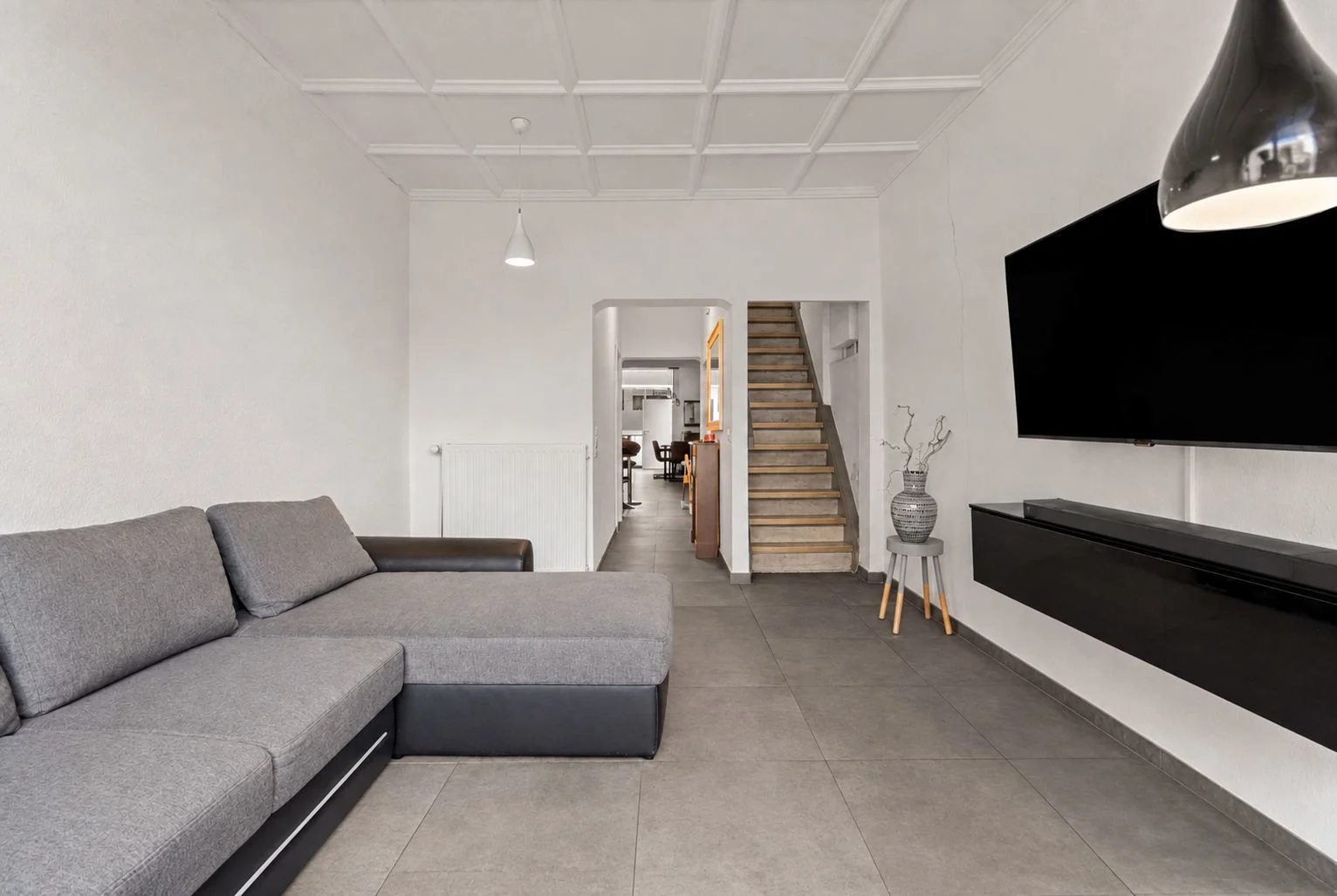 Modernes Wohnzimmer mit grauem Ecksofa, großem Fernseher und Treppe im Hintergrund.