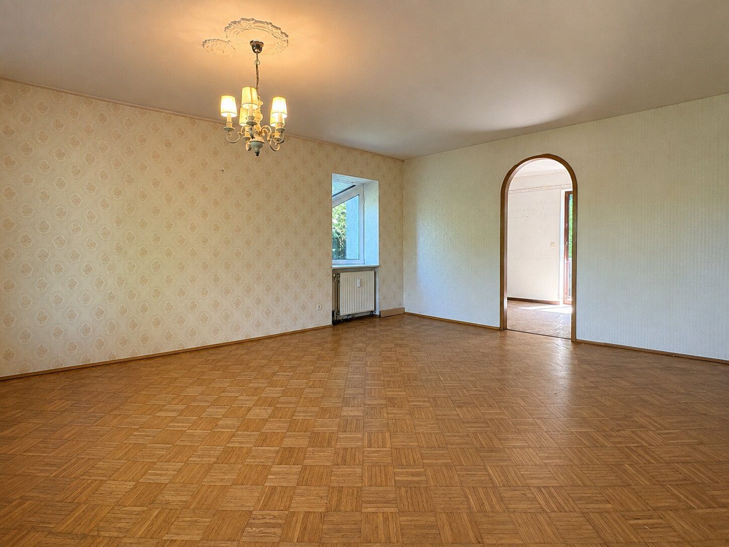 Leeres Wohnzimmer mit Parkettboden, Kronleuchter, Fenster und Rundbogen zur nächsten Tür.