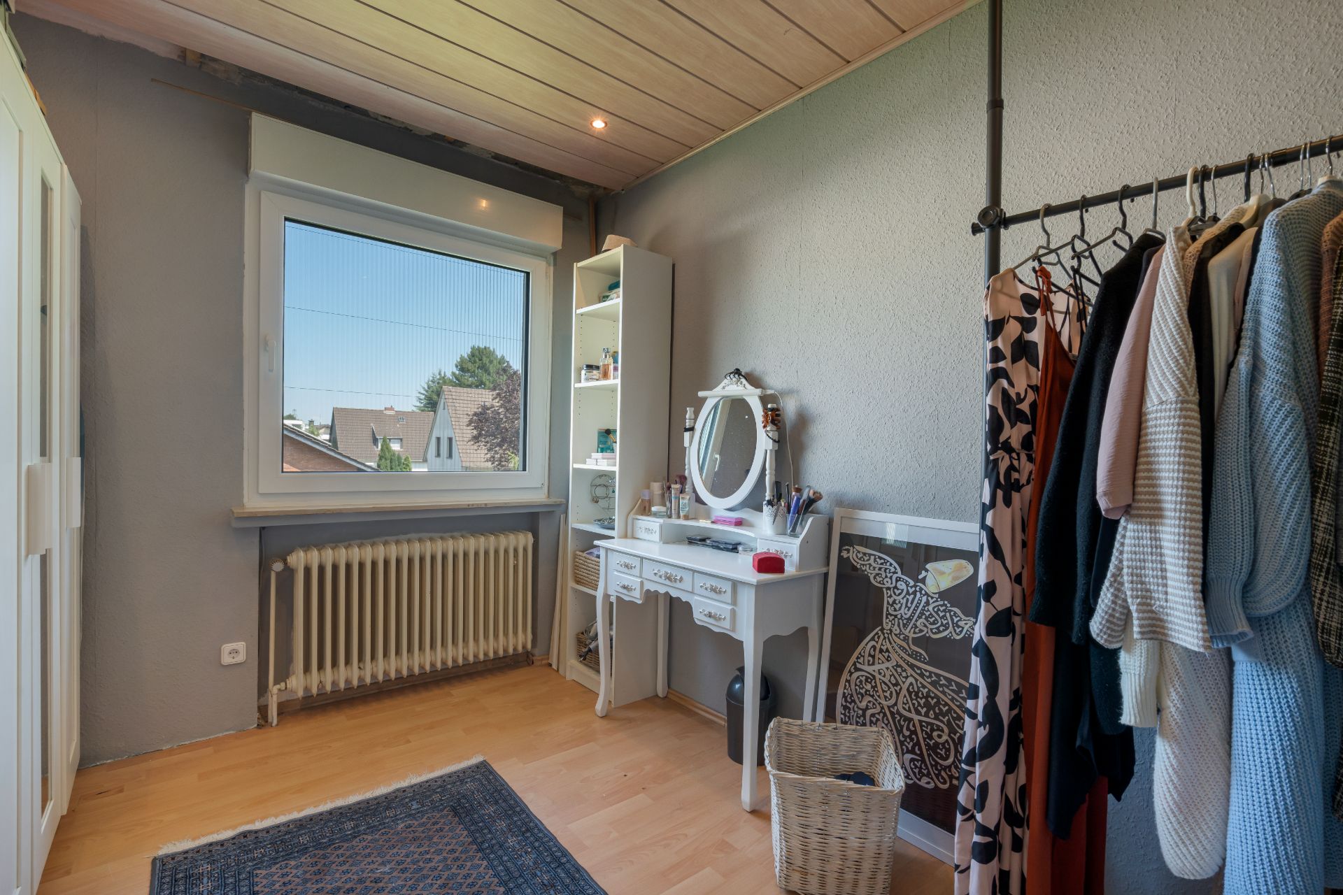 Helles Zimmer mit Schminktisch, Kleiderständer und Regal, großes Fenster mit Blick nach draußen.