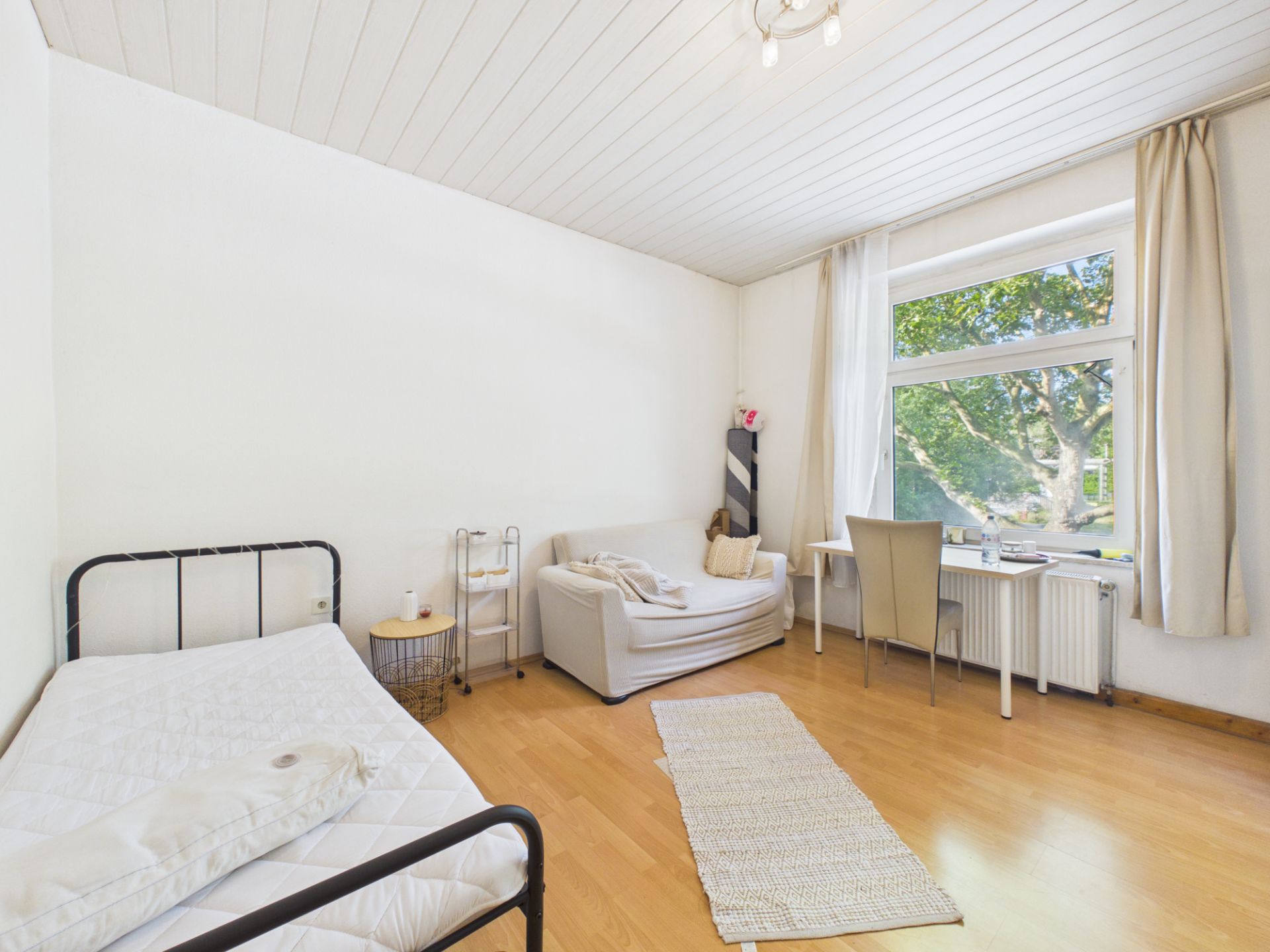 Helles Zimmer mit Bett, Sofa, Schreibtisch und großem Fenster mit Blick auf Bäume.