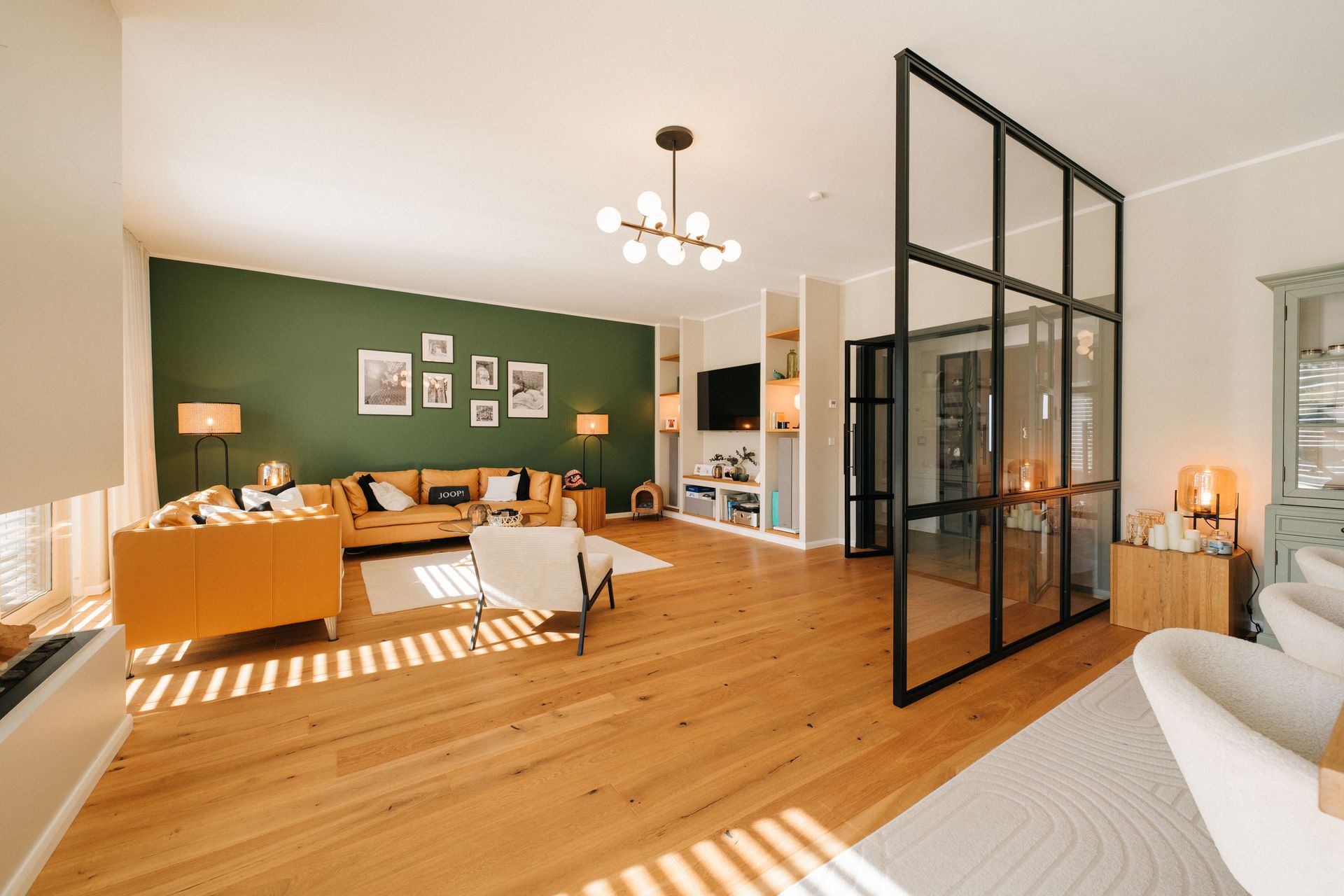 Helles Wohnzimmer mit Holzboden, grüner Wand, Sofa, Sesseln und Glastrennwand.