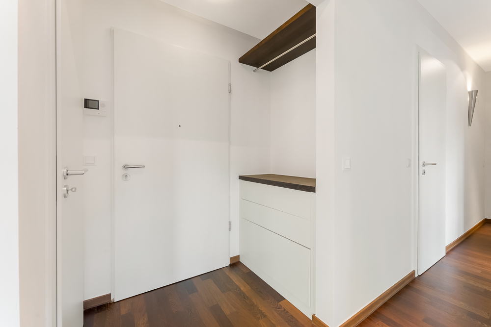 Heller Flur mit Holzboden, weißer Eingangstür und Kommode mit dunkler Ablage.