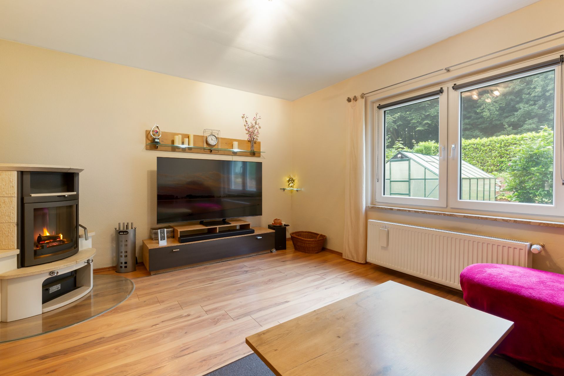 Wohnzimmer mit Fernseher, Kamin, Holzboden und zwei Fenstern mit Blick auf Garten.