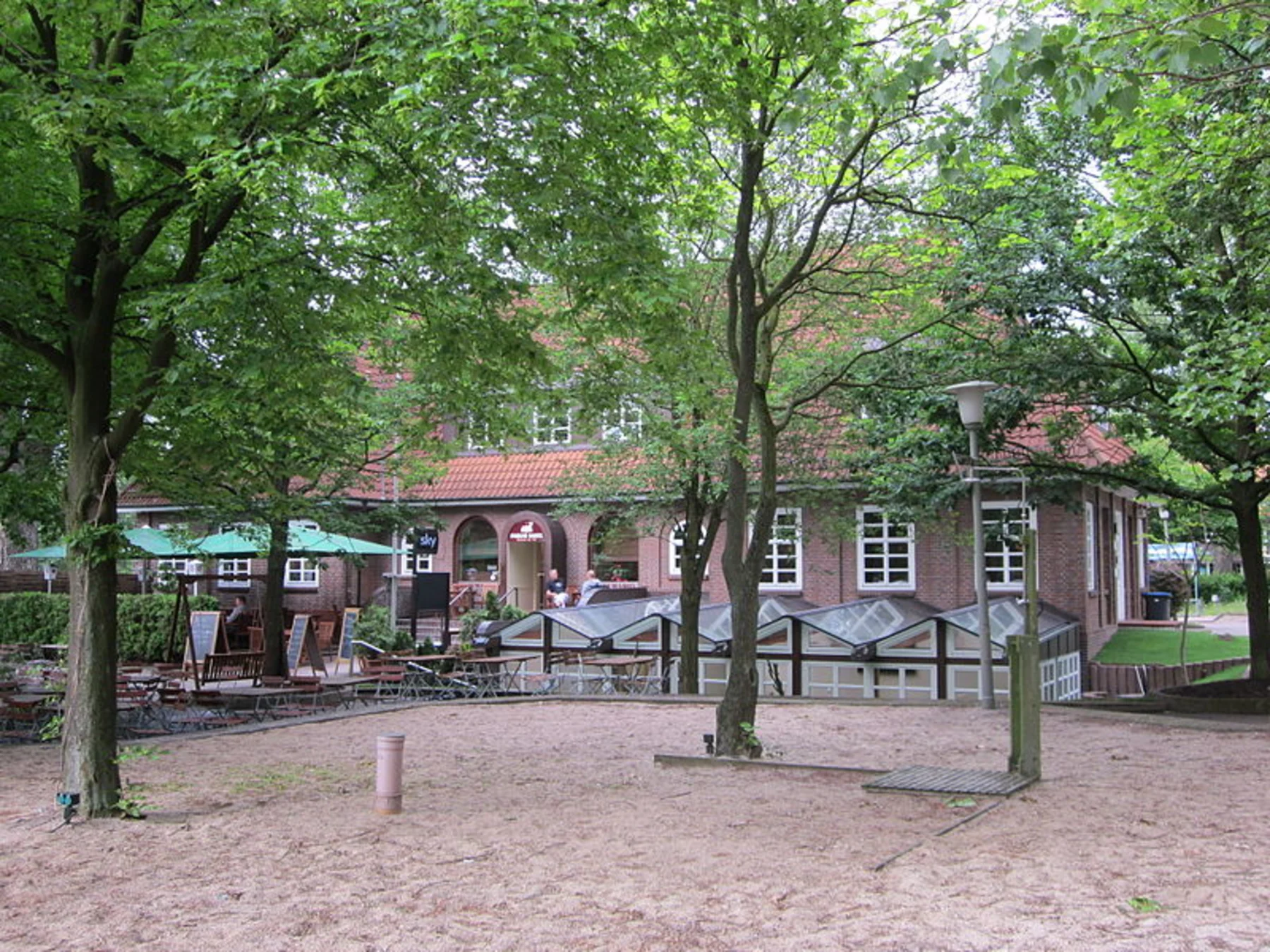 Backsteinwohnhaus mit roten Dachziegeln, umgeben von Bäumen und einem sandigen Vorplatz.