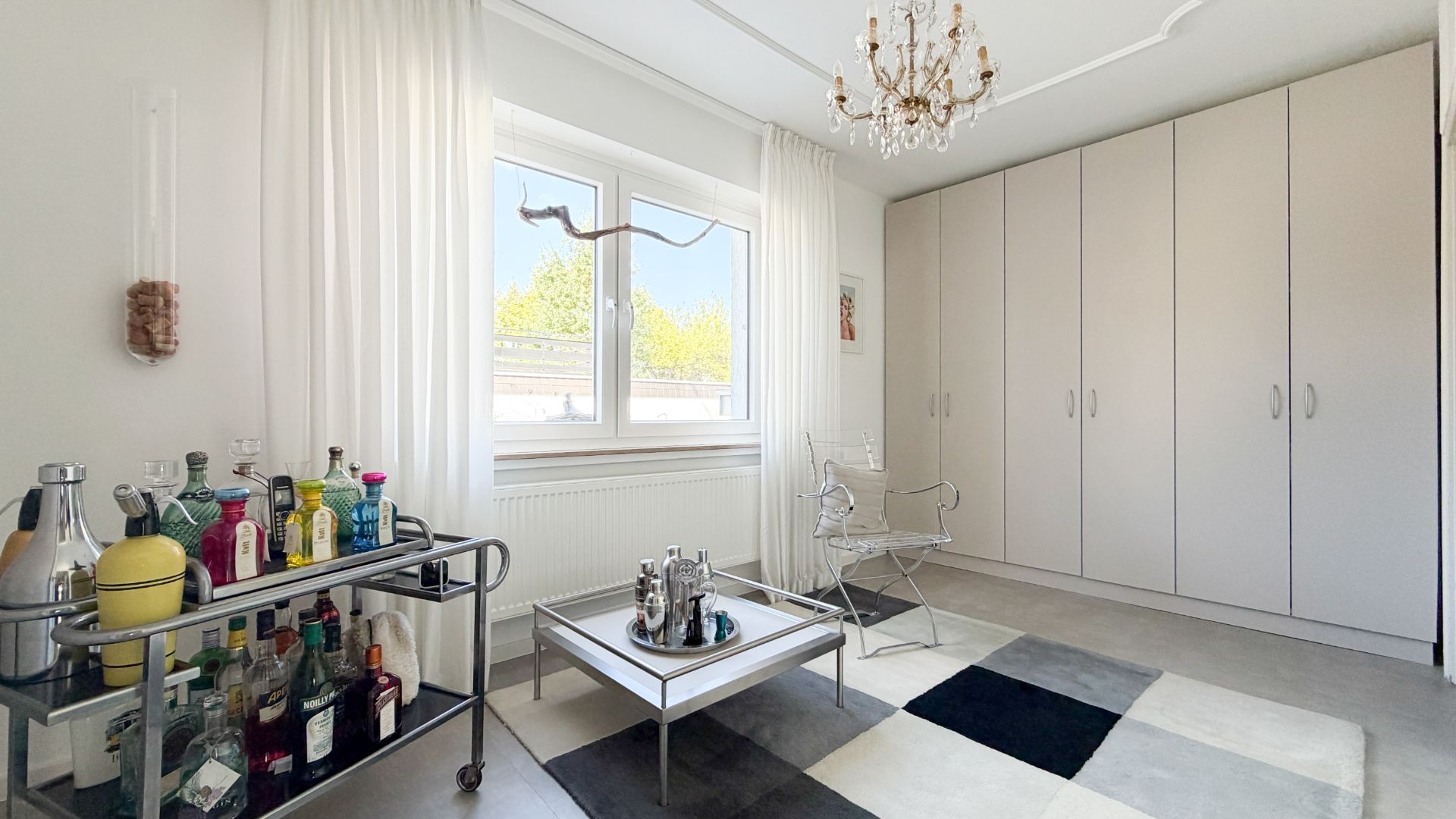 Helles Zimmer mit Minibar, Couchtisch, zwei Stühlen, großem Schrank und Fenster mit weißen Vorhängen.