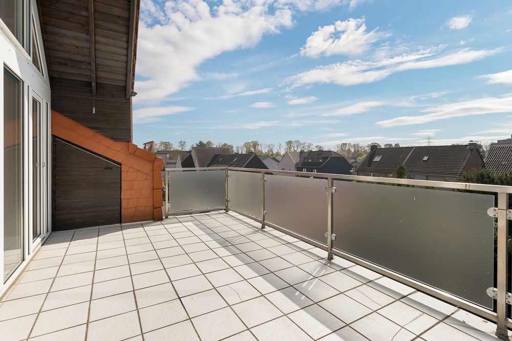 Großer Balkon mit Fliesenboden und Glasgeländer, Blick auf Nachbarhäuser und Himmel.
