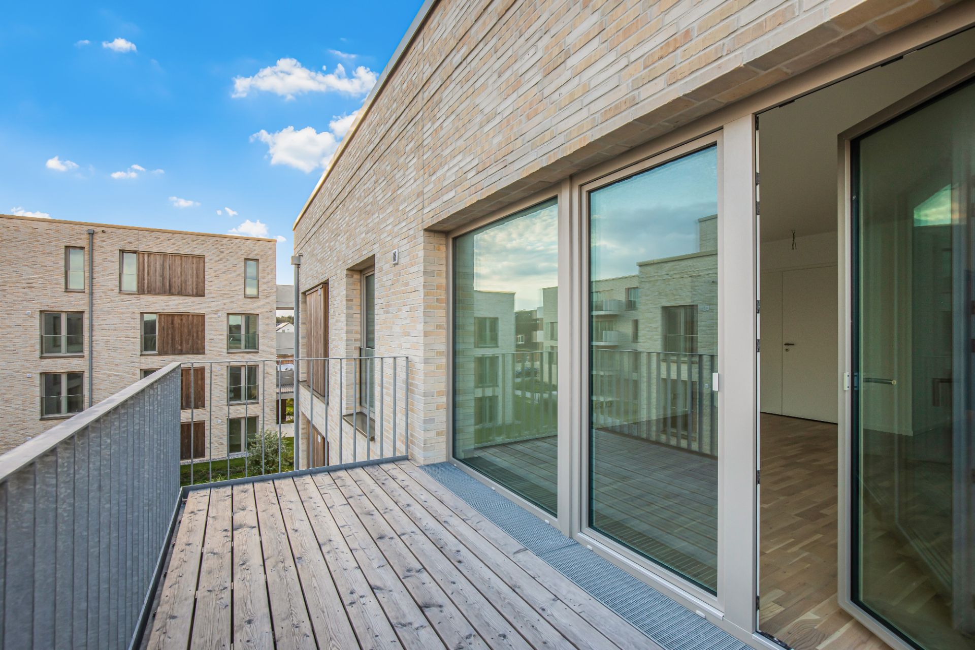 Holzbalkon mit Glas-Schiebetüren, Blick auf moderne Wohngebäude bei blauem Himmel.