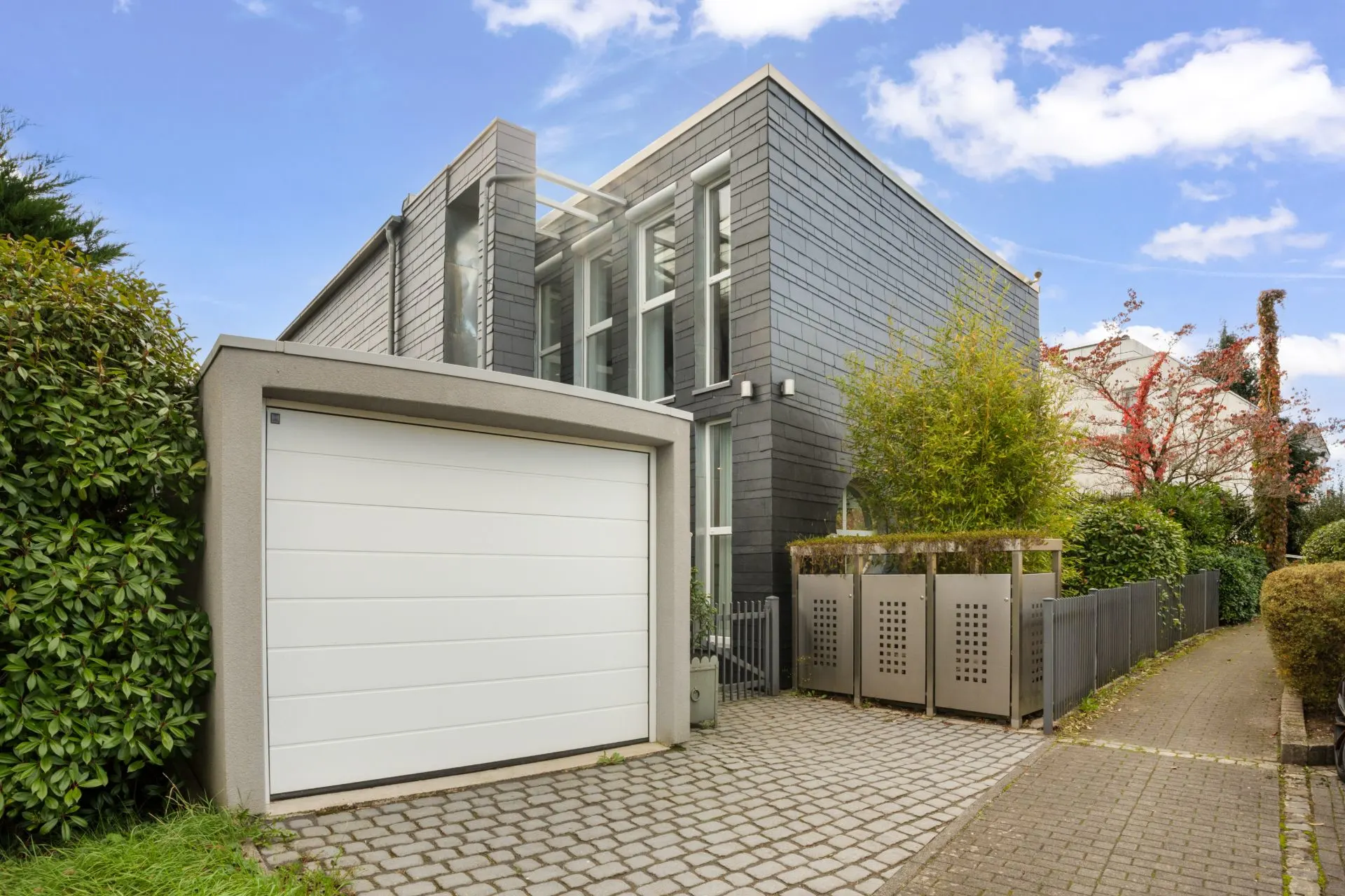 Modernes Wohnhaus mit grauer Fassade, großem Fenster und Garage, umgeben von Pflanzen.