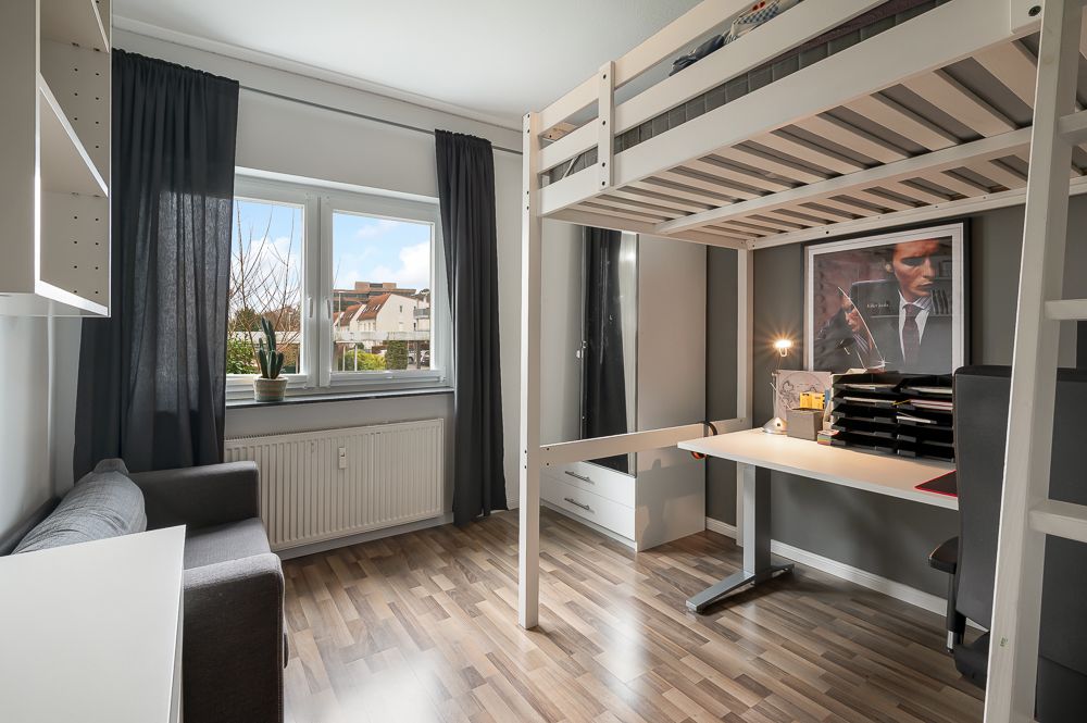 Modernes Zimmer mit Hochbett, Schreibtisch, grauem Sofa und großem Fenster mit dunklen Vorhängen.