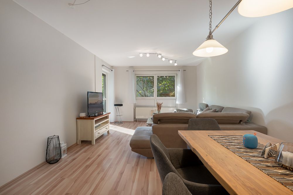 Helles Wohnzimmer mit Holztisch, grauem Sofa, Fernseher und großem Fenster mit Blick ins Grüne.