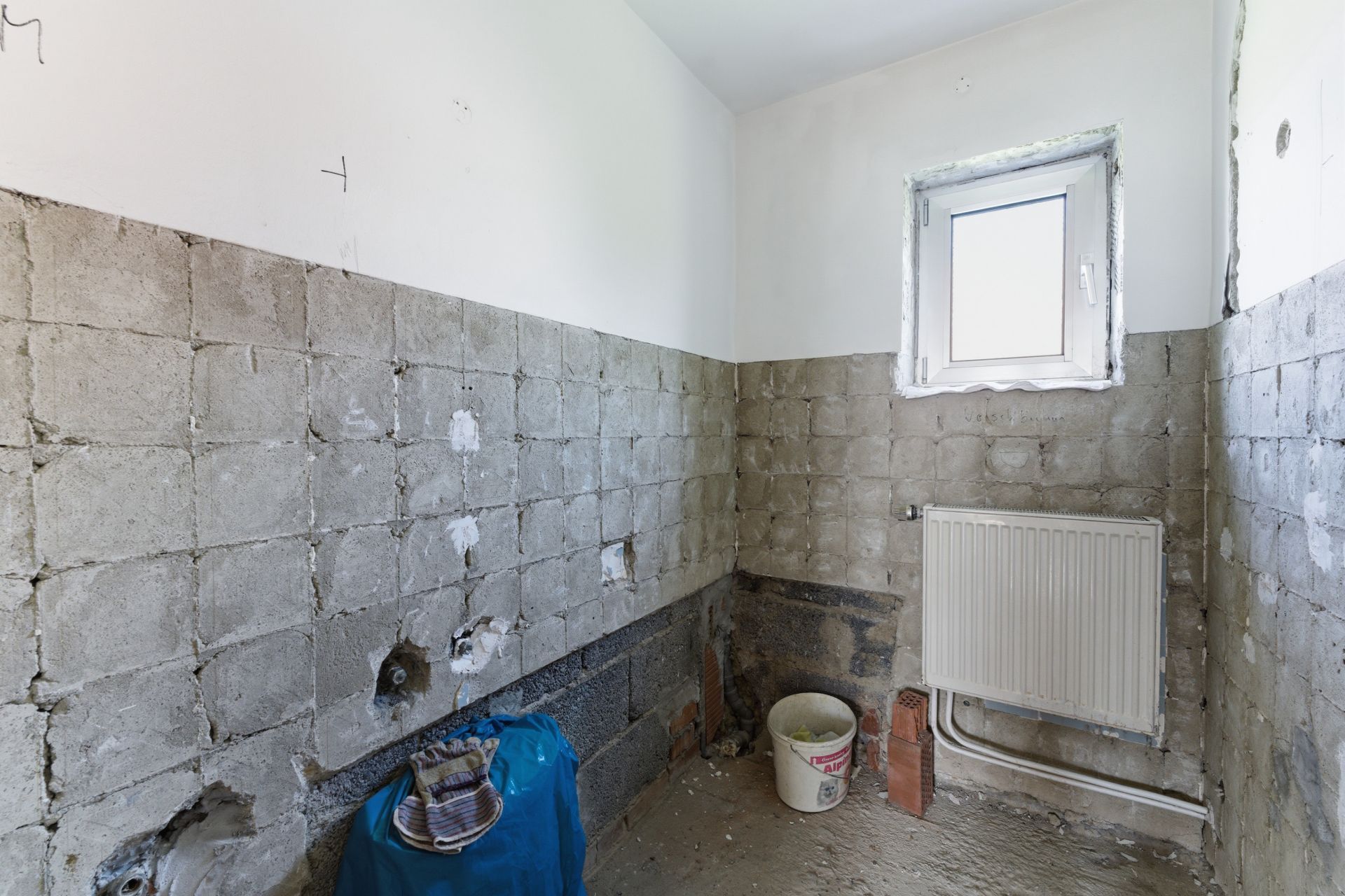 Kleiner Raum mit unverputzten Wänden, Fenster, Heizung und Renovierungsbedarf.