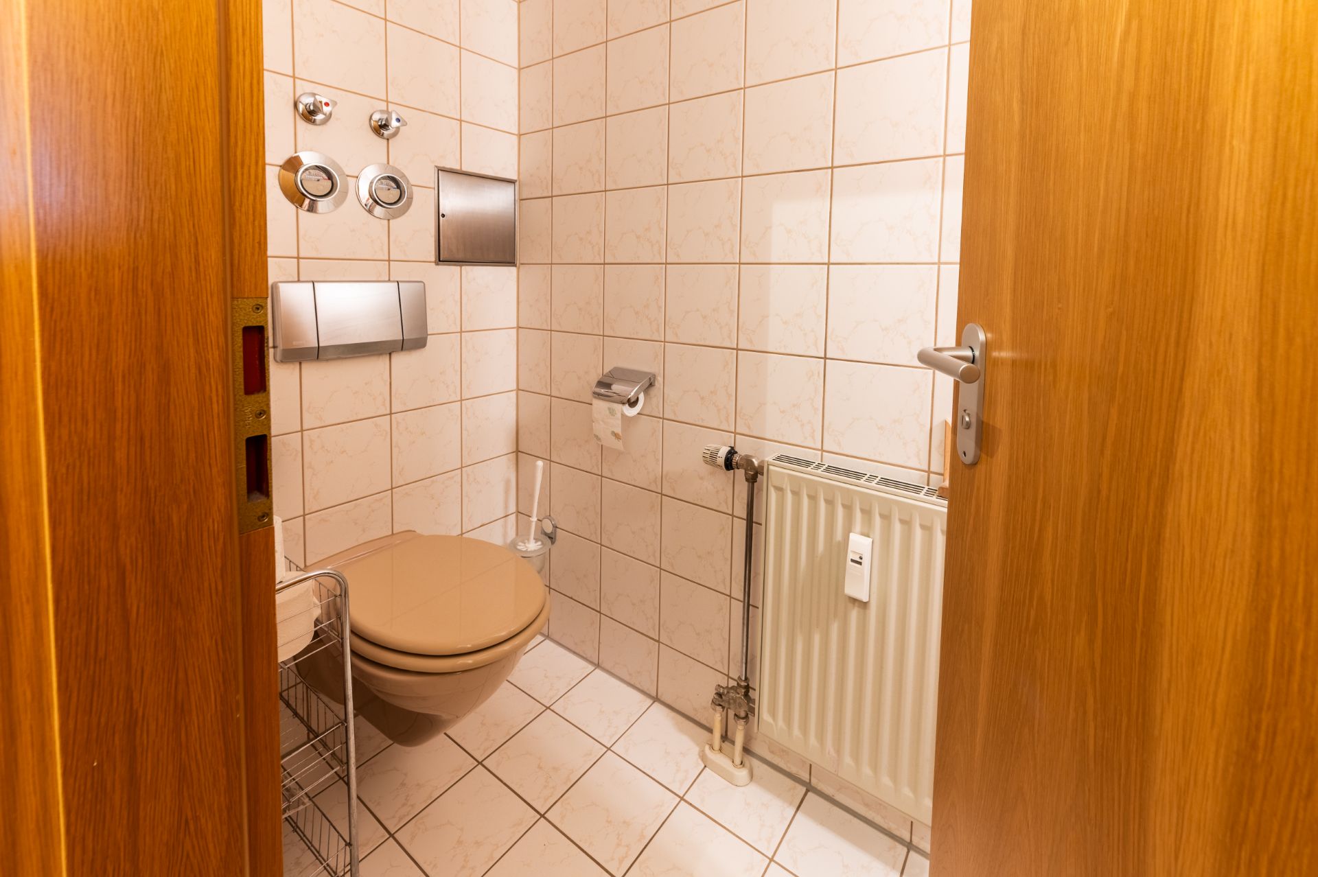 Kleines Badezimmer mit Wandtoilette, Heizkörper, Toilettenpapierhalter und beigen Fliesen.