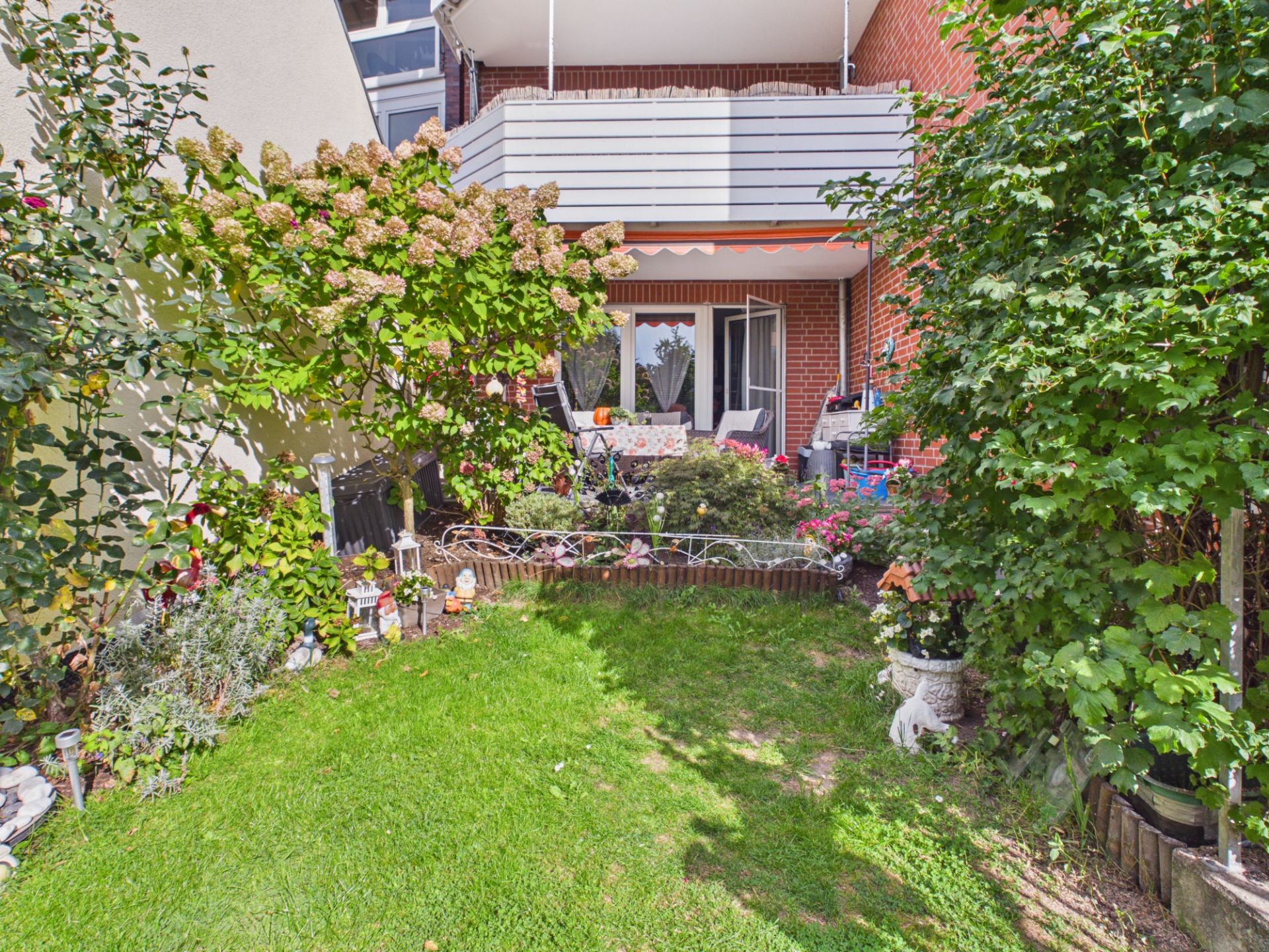Kleiner Garten mit Terrasse, vielen Pflanzen und Sitzmöbeln vor einem Wohnhaus.