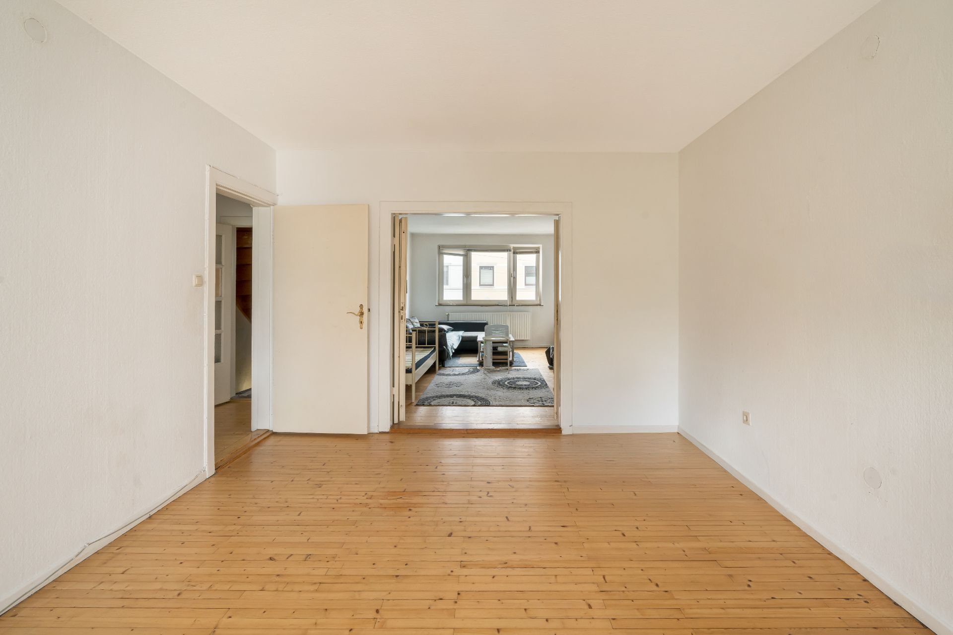 Leerer Raum mit Holzboden, offener Tür und Blick in ein Wohnzimmer mit Sofa und Teppich.