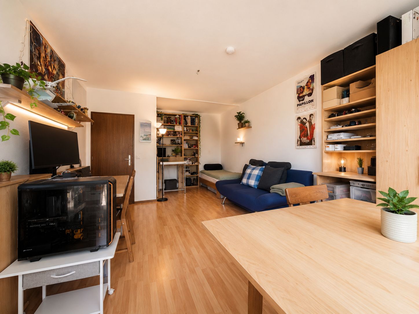 Helles Wohnzimmer mit Sofa, Schreibtisch, Regalen, Computer und Holztisch, Pflanzen und Dekoration.