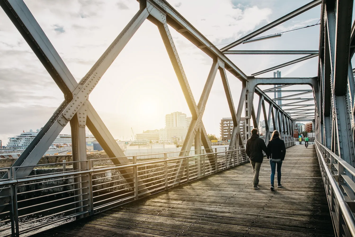 Zwei Personen gehen auf einer Metallbrücke mit Holzbohlen, Stadtgebäude im Hintergrund.