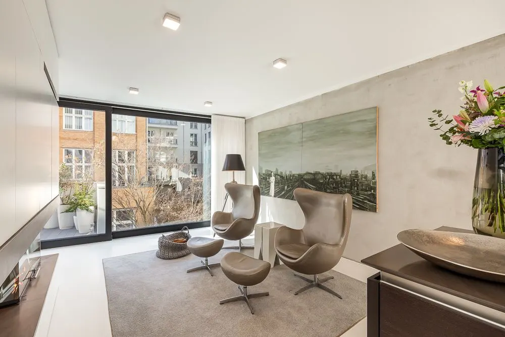 Modernes Wohnzimmer mit großen Fenstern, zwei Sesseln, Hocker, Teppich und Blumenstrauß.
