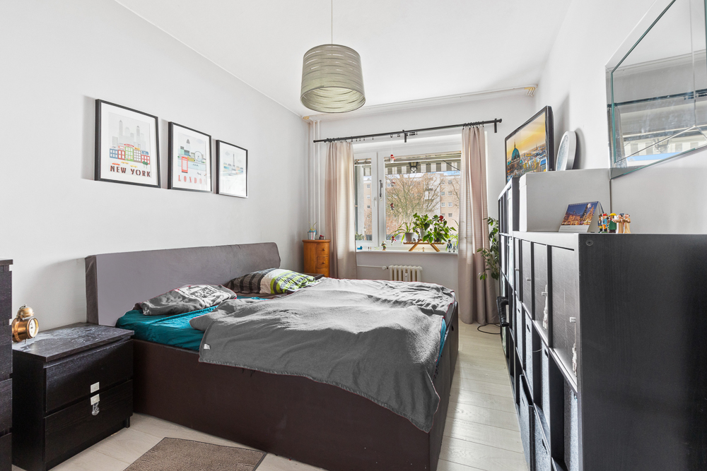 Helles Schlafzimmer mit Doppelbett, Wandbildern, Regal und großem Fenster mit Vorhängen.