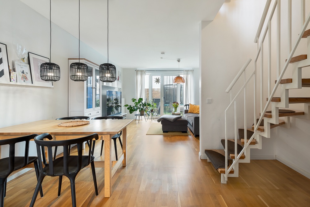 Helles Wohnzimmer mit Esstisch, schwarzen Stühlen, Sofa, Pflanzen und Treppe.