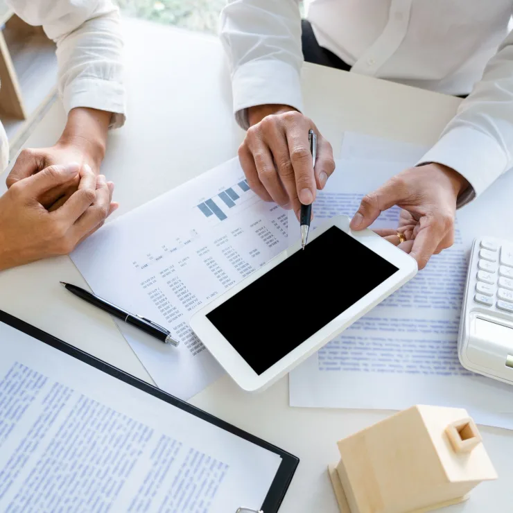 Zwei Personen besprechen Dokumente mit Tablet, Taschenrechner und kleinem Modellhaus auf dem Tisch.