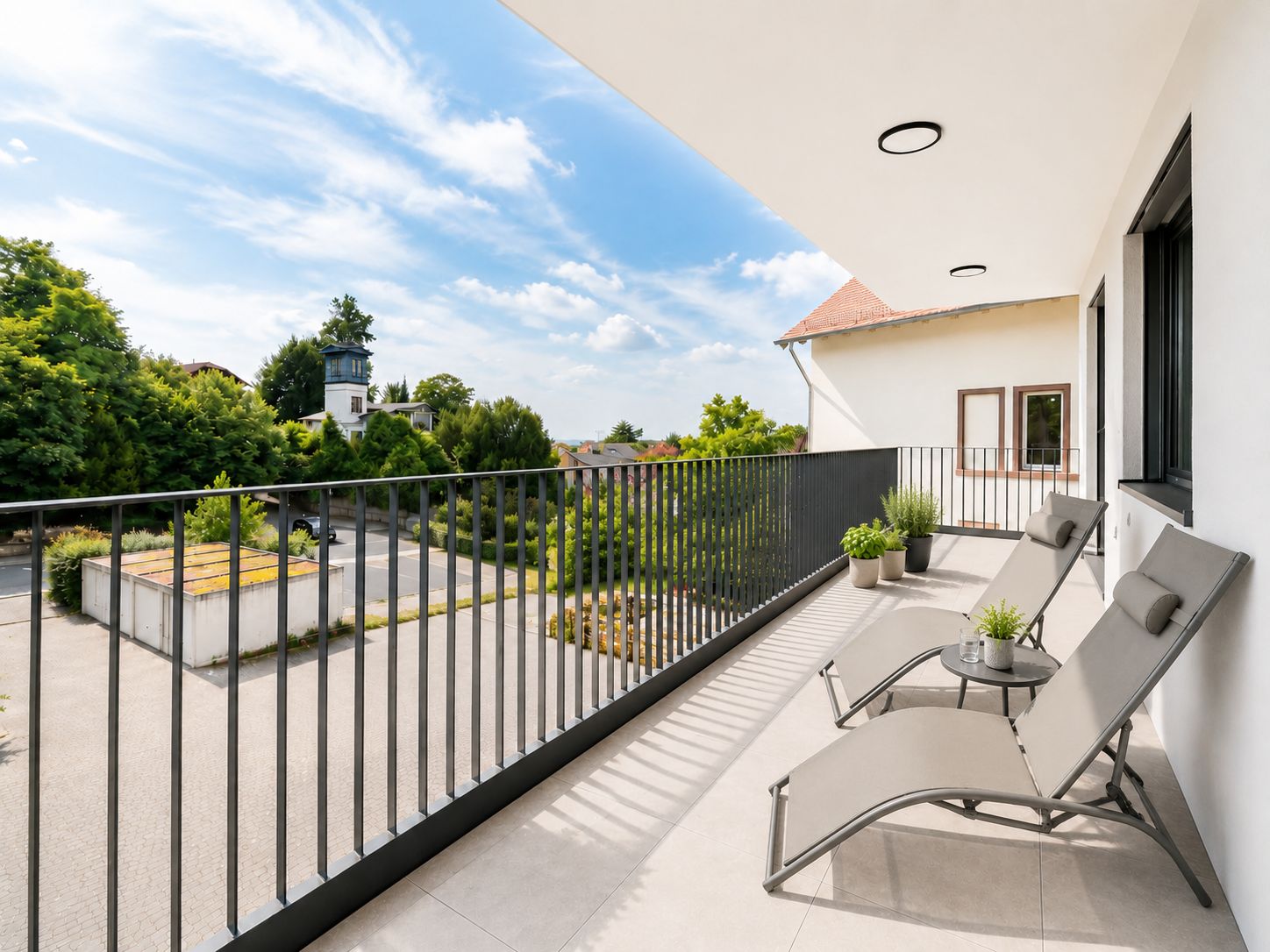 Großer Balkon mit zwei Liegestühlen, kleinen Tischen und Pflanzen, Blick auf grüne Bäume.