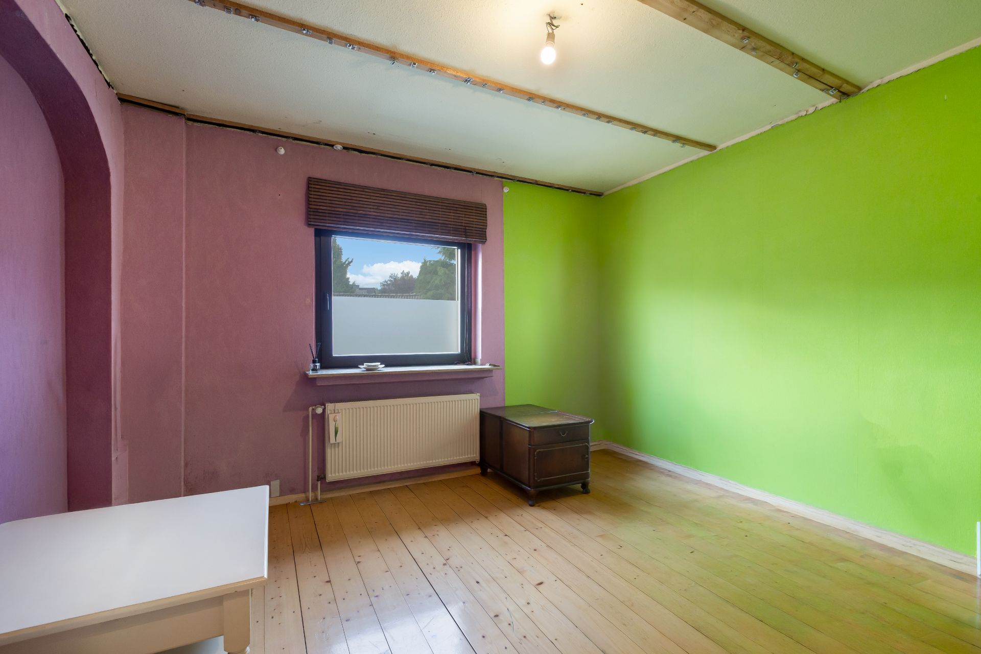 Zimmer mit grün und lila Wänden, Holzboden, Fenster, Tisch und Kommode.