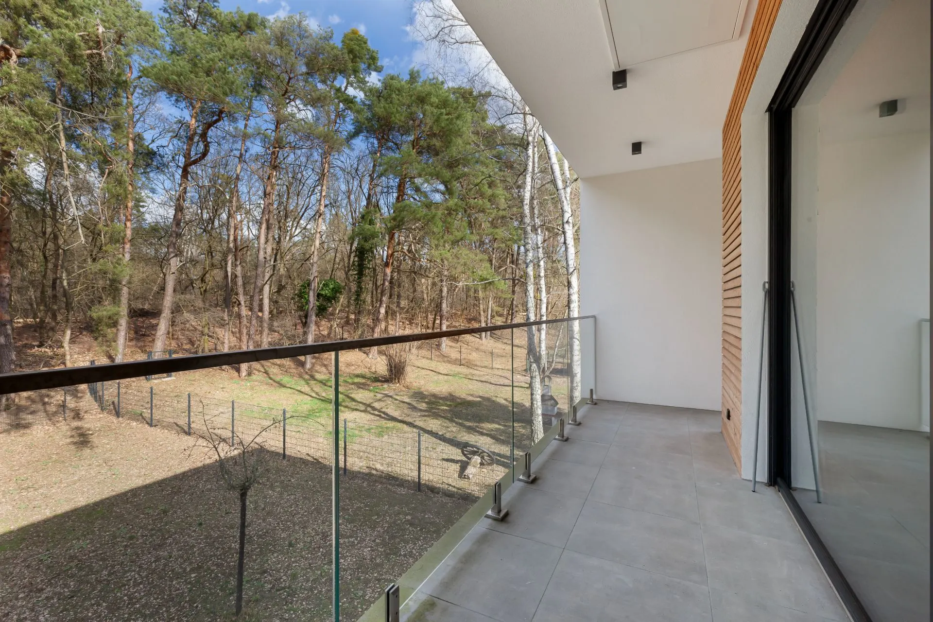 Moderner Balkon mit Glasgeländer, Blick auf einen Garten mit Bäumen.