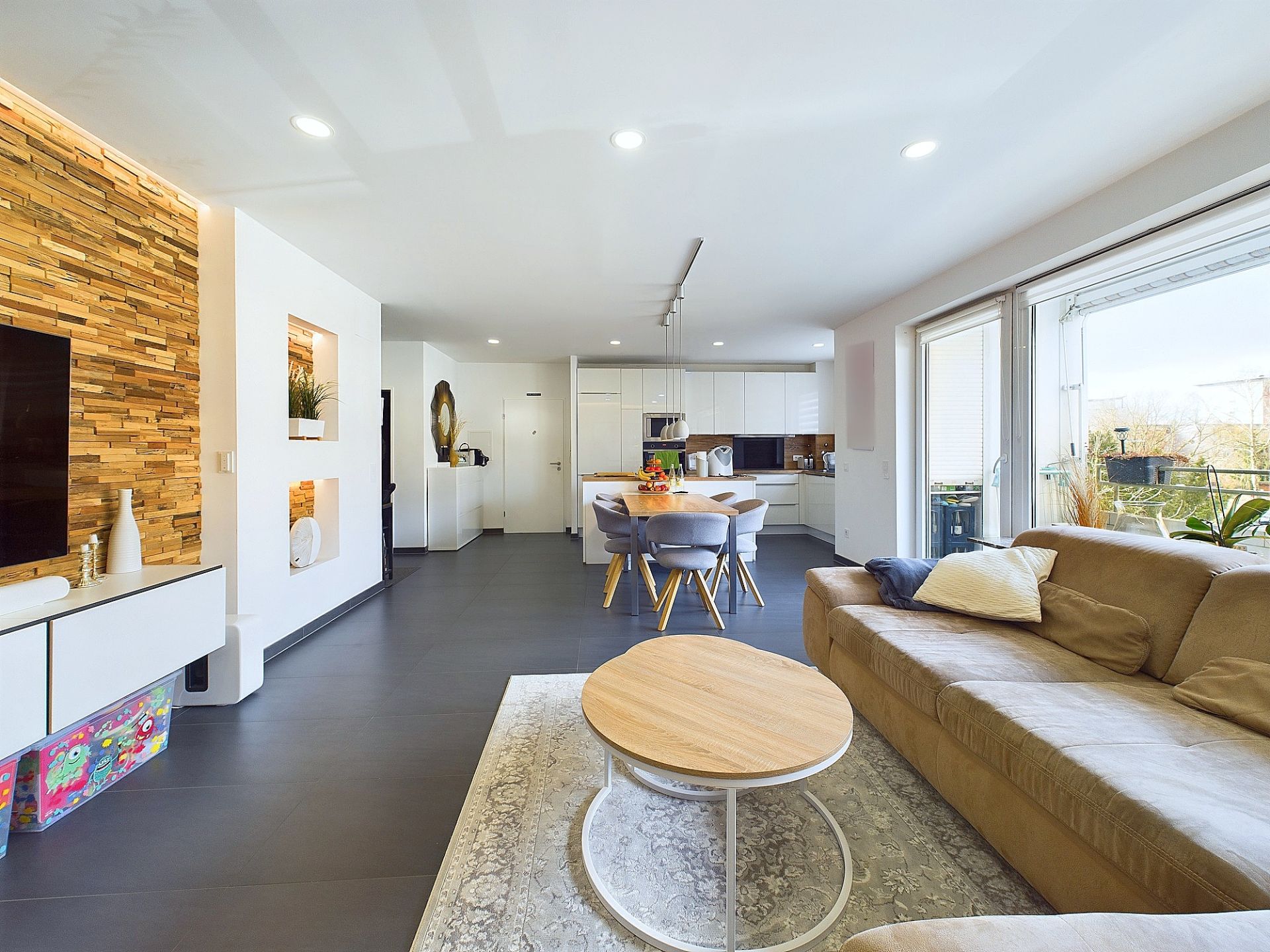 Modernes Wohnzimmer mit Sofa, Couchtisch, Esstisch, offener Küche und großen Fenstern zum Balkon.